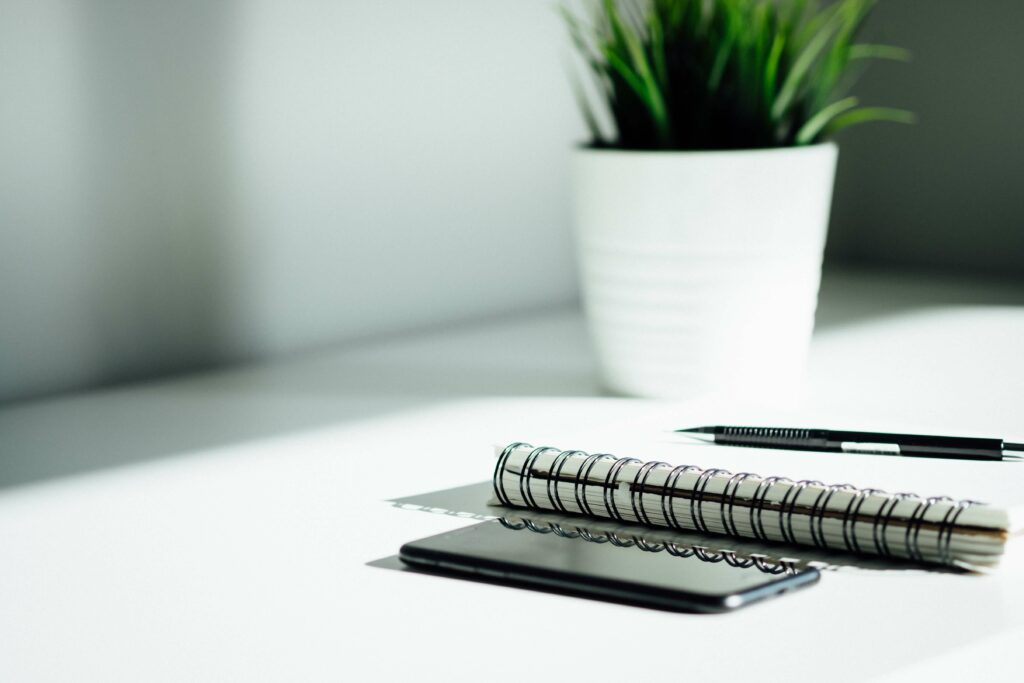 Auf dem Tisch liegen Handy, Block und Stift nebeneinander. Im Hintergrund ist eine Pflanze